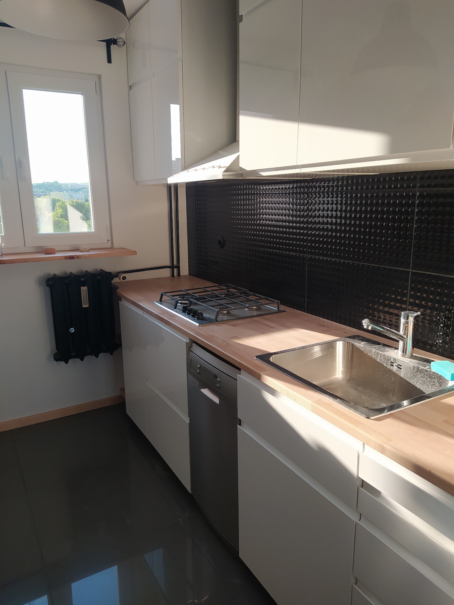 Nowoczesna kuchnia z białymi szafkami, bukowym blatem, czarną, wypukłą mozaiką na ścianie, widokiem z okna i starodawnym grzejnikiem żeliwnym.