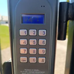 Panel domofonu z klawiaturą numeryczną i wyświetlaczem LCD prezentującym numer lokalu, zamontowany na metalowym słupku bramy.