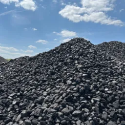 Duży pryzmat czarnych bryłek węgla na tle błękitnego nieba z białymi obłokami.