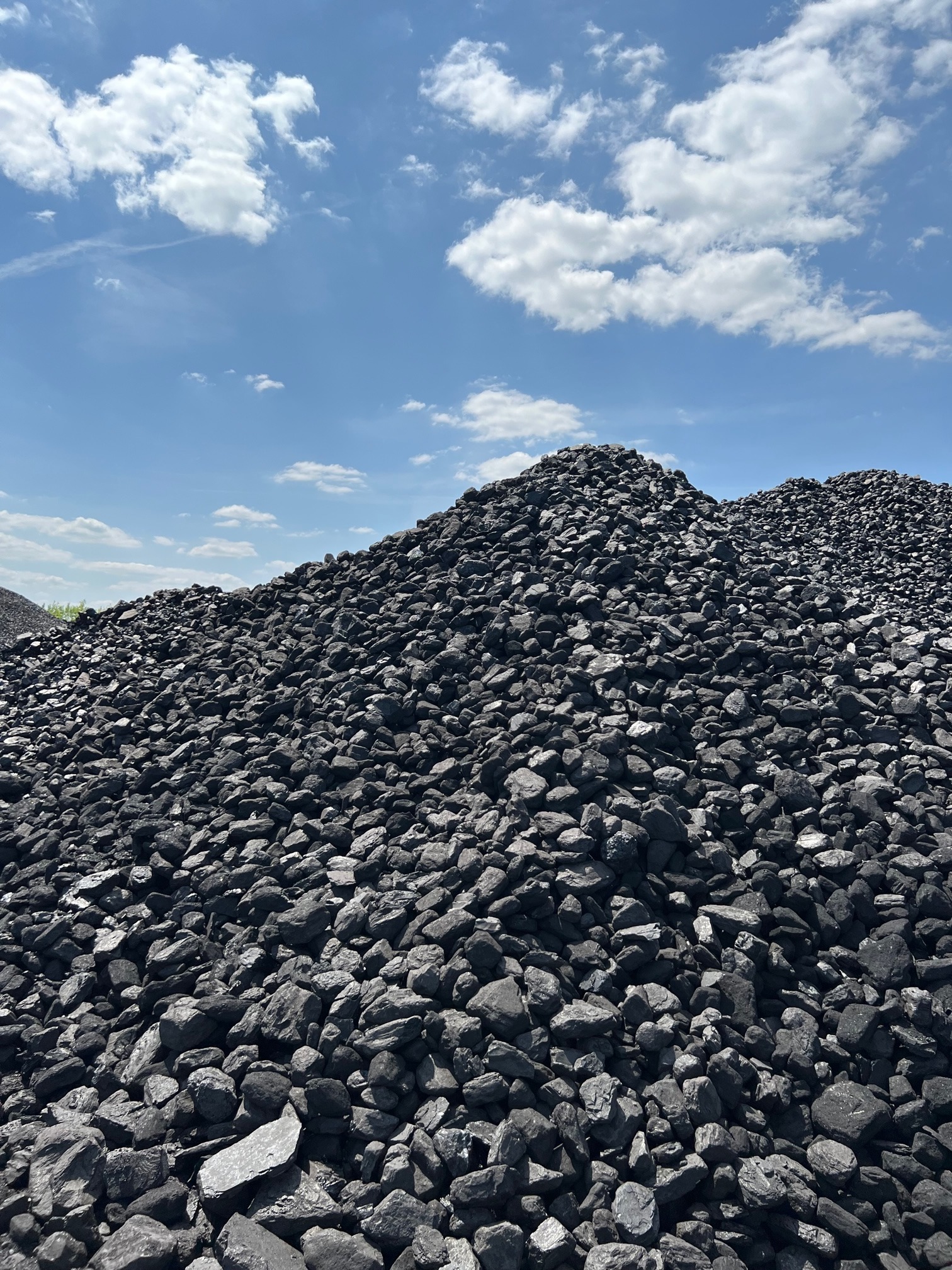 Duży pryzmat czarnych bryłek węgla na tle błękitnego nieba z białymi obłokami.