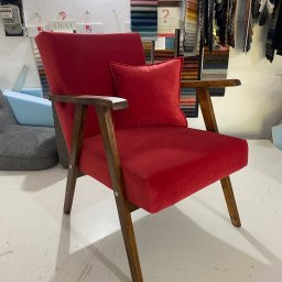 Mateusz Kowalski - Czerwony, tapicerowany fotel z drewnianymi podłokietnikami i poduszką, prezentowany w pracowni z próbkami tkanin w tle.