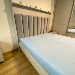 Mateusz Kowalski - Nowoczesne łóżko z tapicerowanym wezgłowiem w odcieniach szarości, z dekoracyjną półką na drobne figurki, ustawione w sypialni z przesuwną szafą.