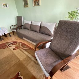 Mateusz Kowalski - Pokój dzienny z brązową sofą z białymi akcentami, dwoma fotelami z drewnianymi podłokietnikami i wzorzystym dywanem na parkiecie, oświetlony naturalnym światłem.