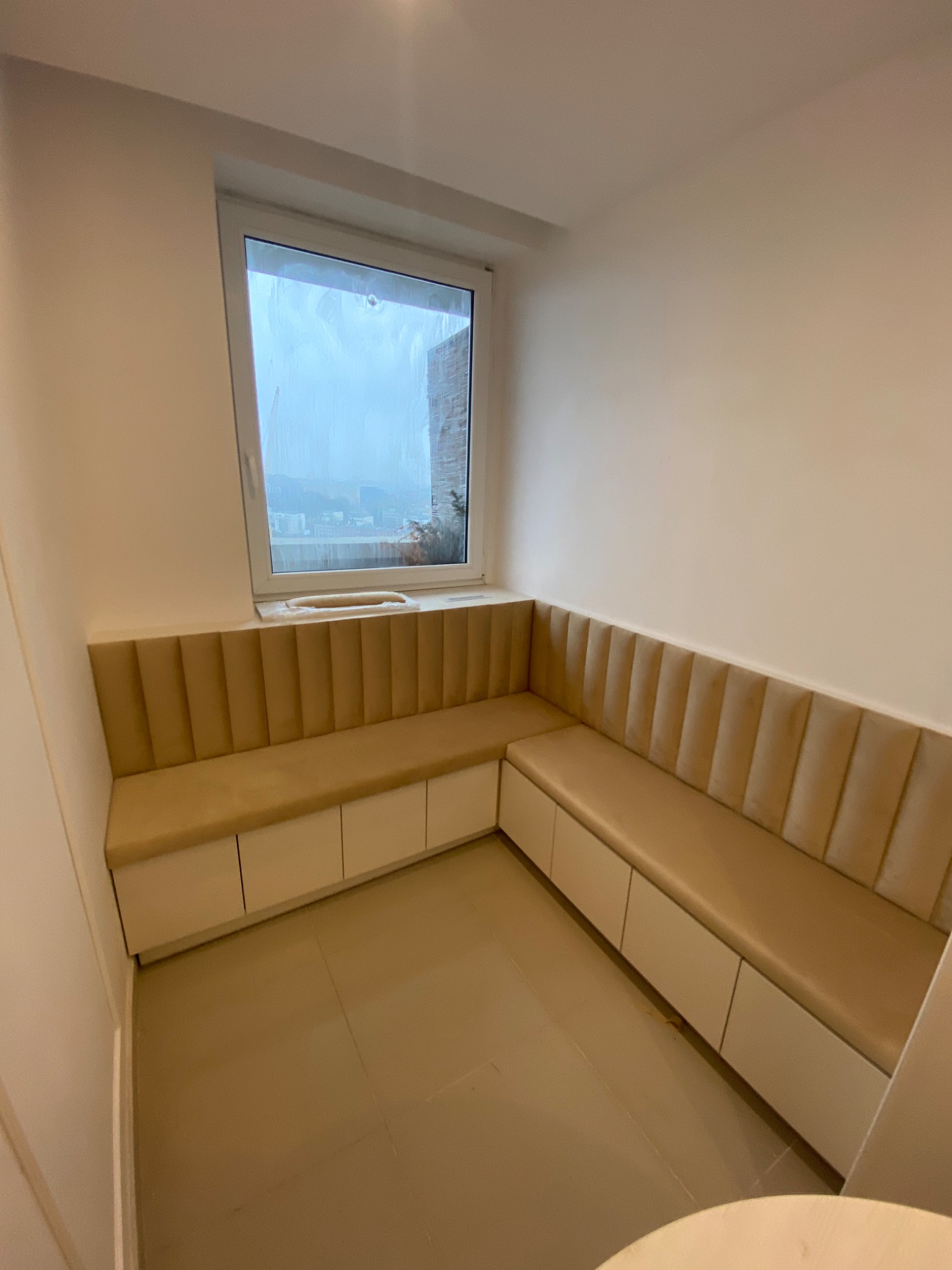 Beżowa, tapicerowana narożna ławka z pojemnikami pod siedziskiem, wnęka okienna z widokiem na miasto w tle.