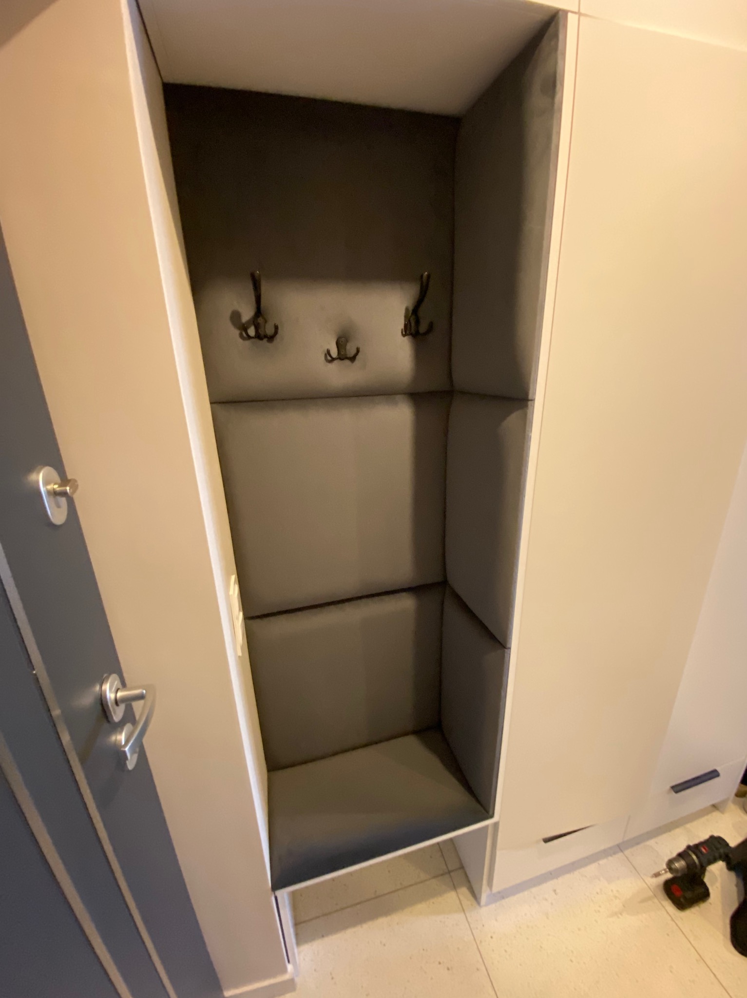 Wnęka w zabudowie meblowej z tapicerowanym siedziskiem i ścianami w szarym kolorze, wyposażona w trzy wieszaki na ubrania.