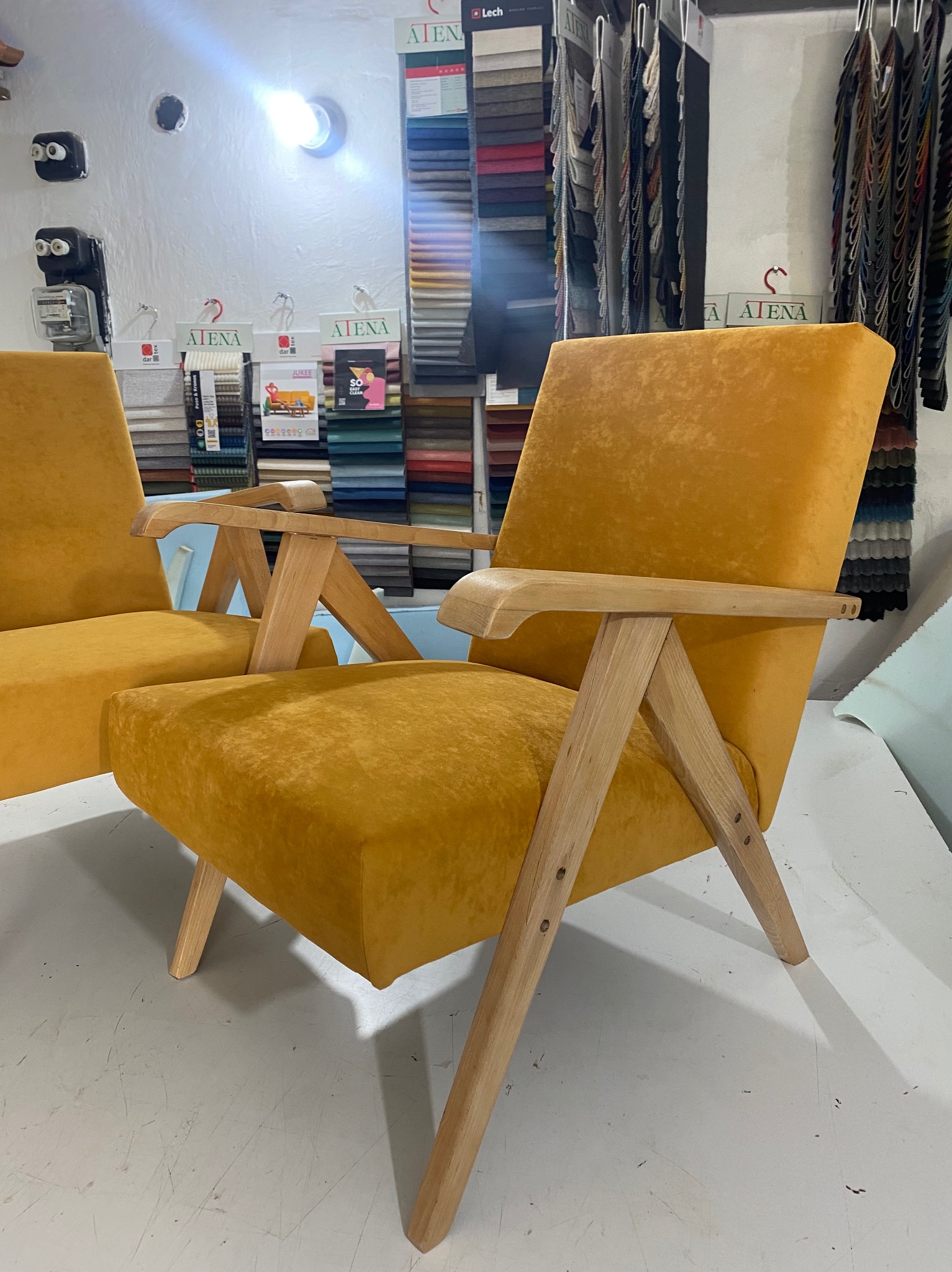 Dwa żółte fotele tapicerowane z drewnianymi podłokietnikami i nogami, w tle ściana z próbkami tkanin.