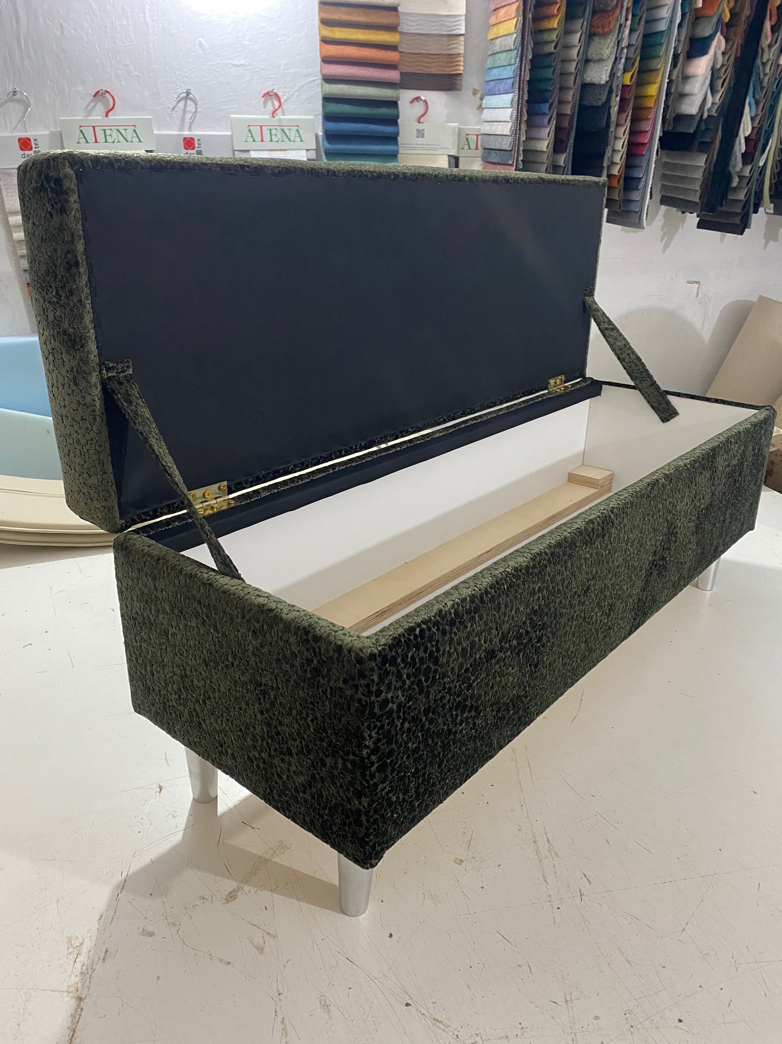 Otwierana pufa tapicerowana zielonym materiałem o wzorzystej fakturze, na srebrnych nóżkach, z widocznym wnętrzem i drewnianymi elementami. W tle wzorniki tkanin.