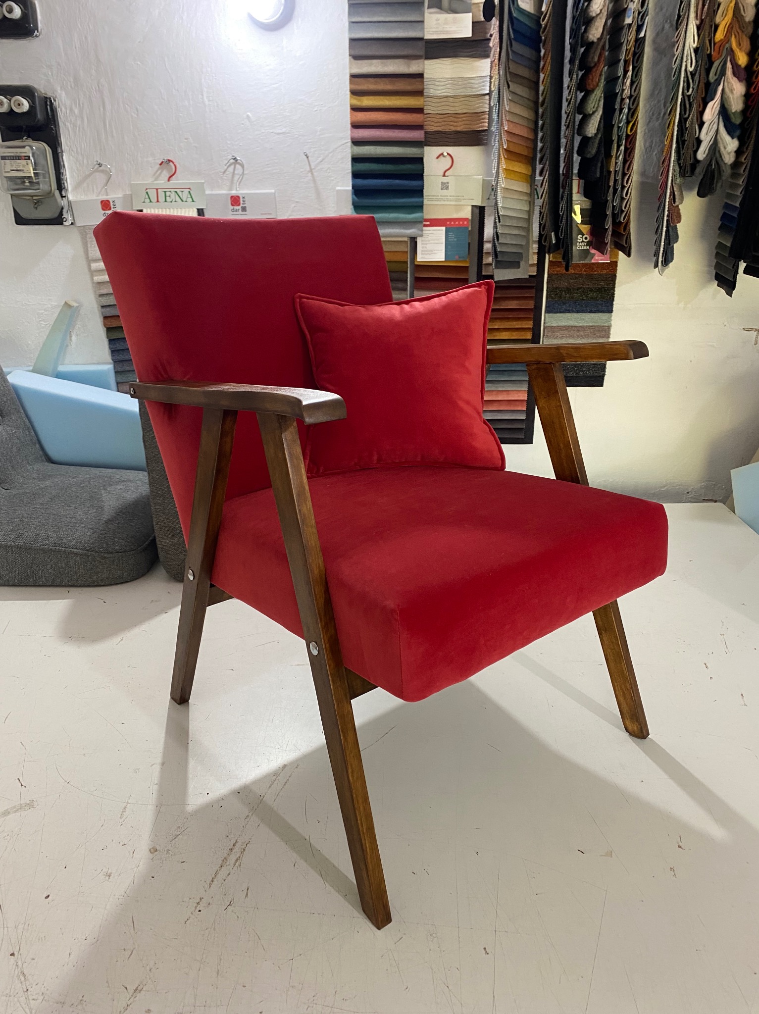 Czerwony, tapicerowany fotel z drewnianymi podłokietnikami i poduszką, prezentowany w pracowni z próbkami tkanin w tle.