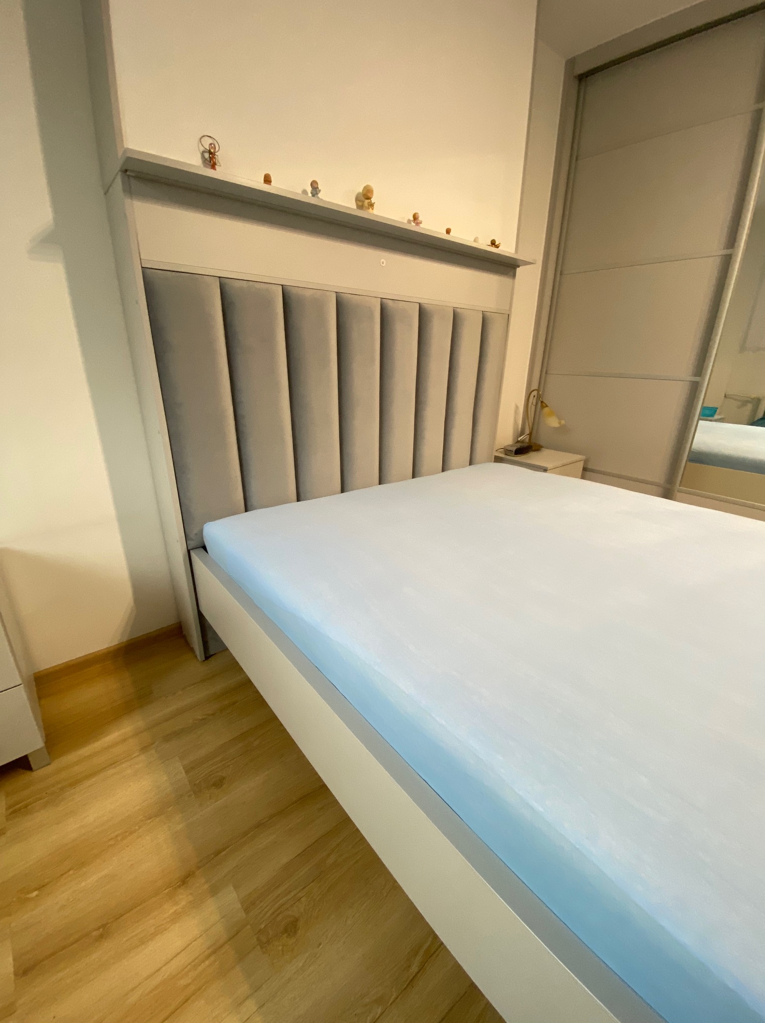 Nowoczesne łóżko z tapicerowanym wezgłowiem w odcieniach szarości, z dekoracyjną półką na drobne figurki, ustawione w sypialni z przesuwną szafą.