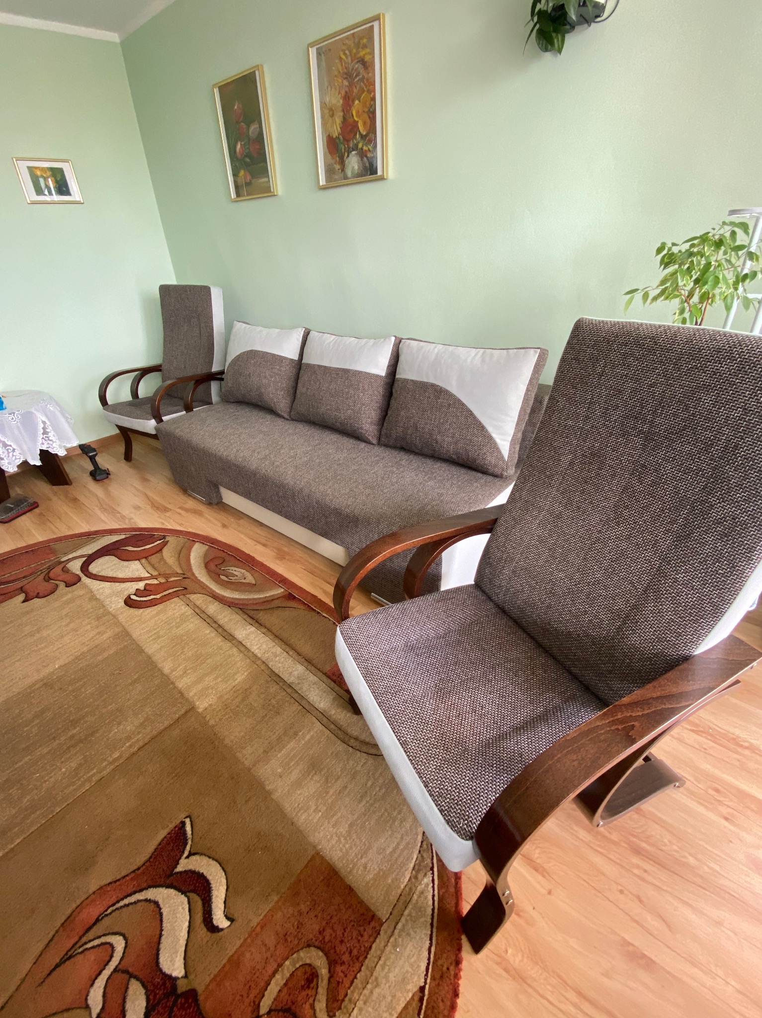 Pokój dzienny z brązową sofą z białymi akcentami, dwoma fotelami z drewnianymi podłokietnikami i wzorzystym dywanem na parkiecie, oświetlony naturalnym światłem.