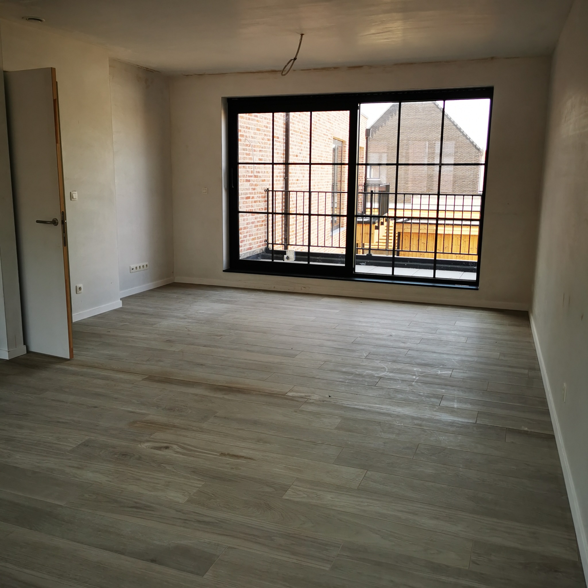 Wnętrze pokoju w trakcie prac wykończeniowych: położona podłoga z płytek imitujących drewno, surowe ściany przygotowane do malowania, widoczna instalacja elektryczna na suficie, okno balkonowe...