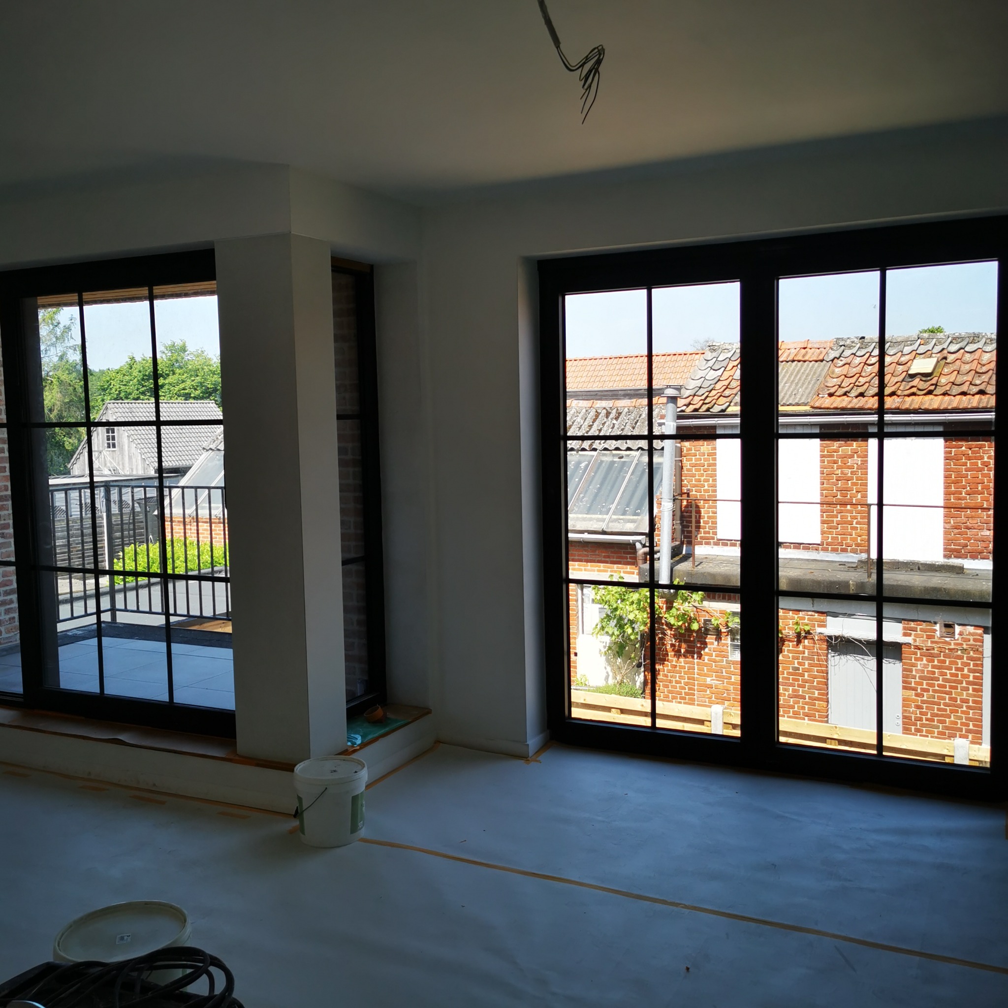 Wnętrze w trakcie prac wykończeniowych, widoczne okna z czarnymi szprosami, zabezpieczona podłoga, ściany pomalowane na biało, wystające kable z sufitu i widok na zewnątrz na zabudowę mieszkalną.
