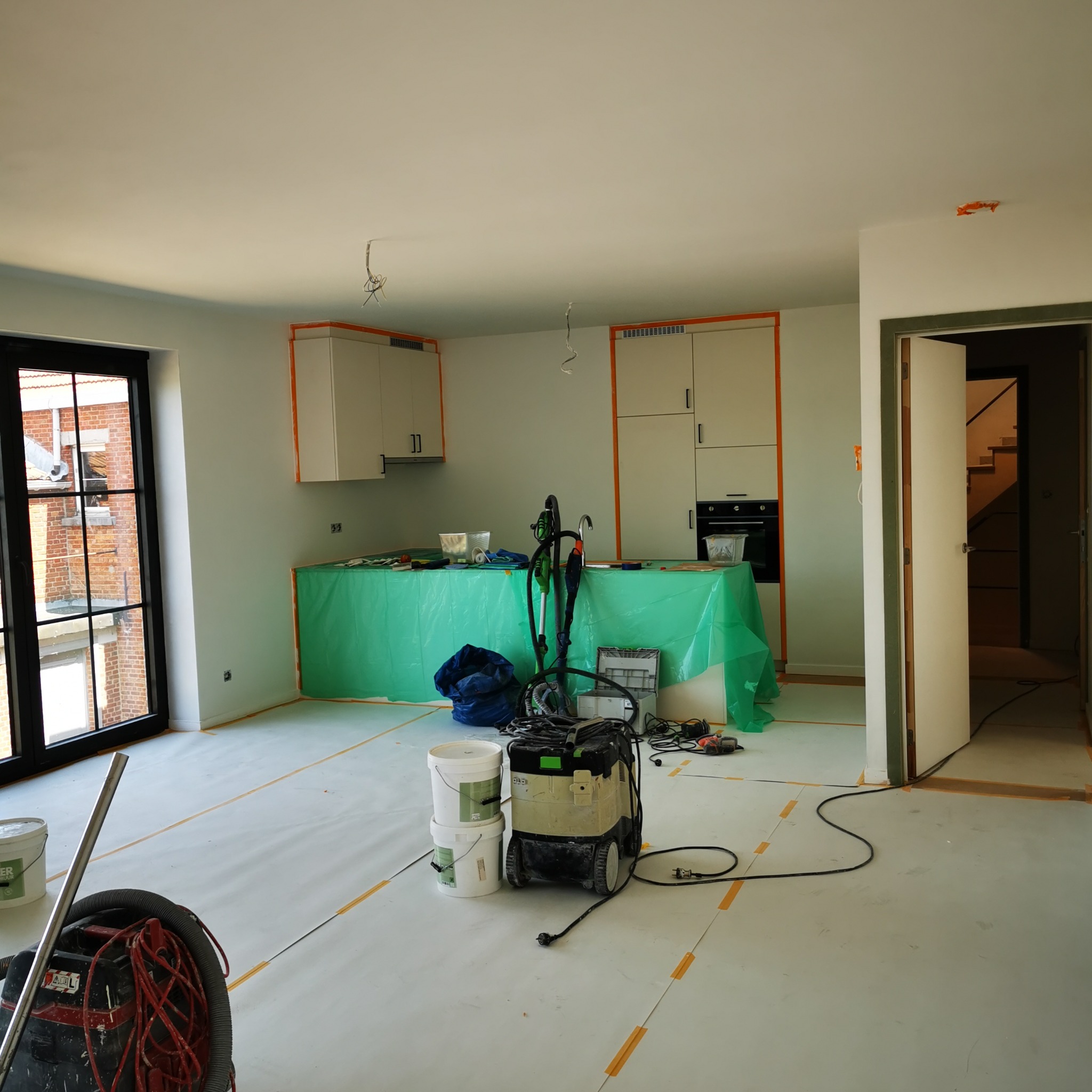 Wnętrze w trakcie prac przygotowawczych: zabezpieczona folią kuchnia, zaklejone taśmą podłogi, widoczne wiadra z farbą i odkurzacz przemysłowy.