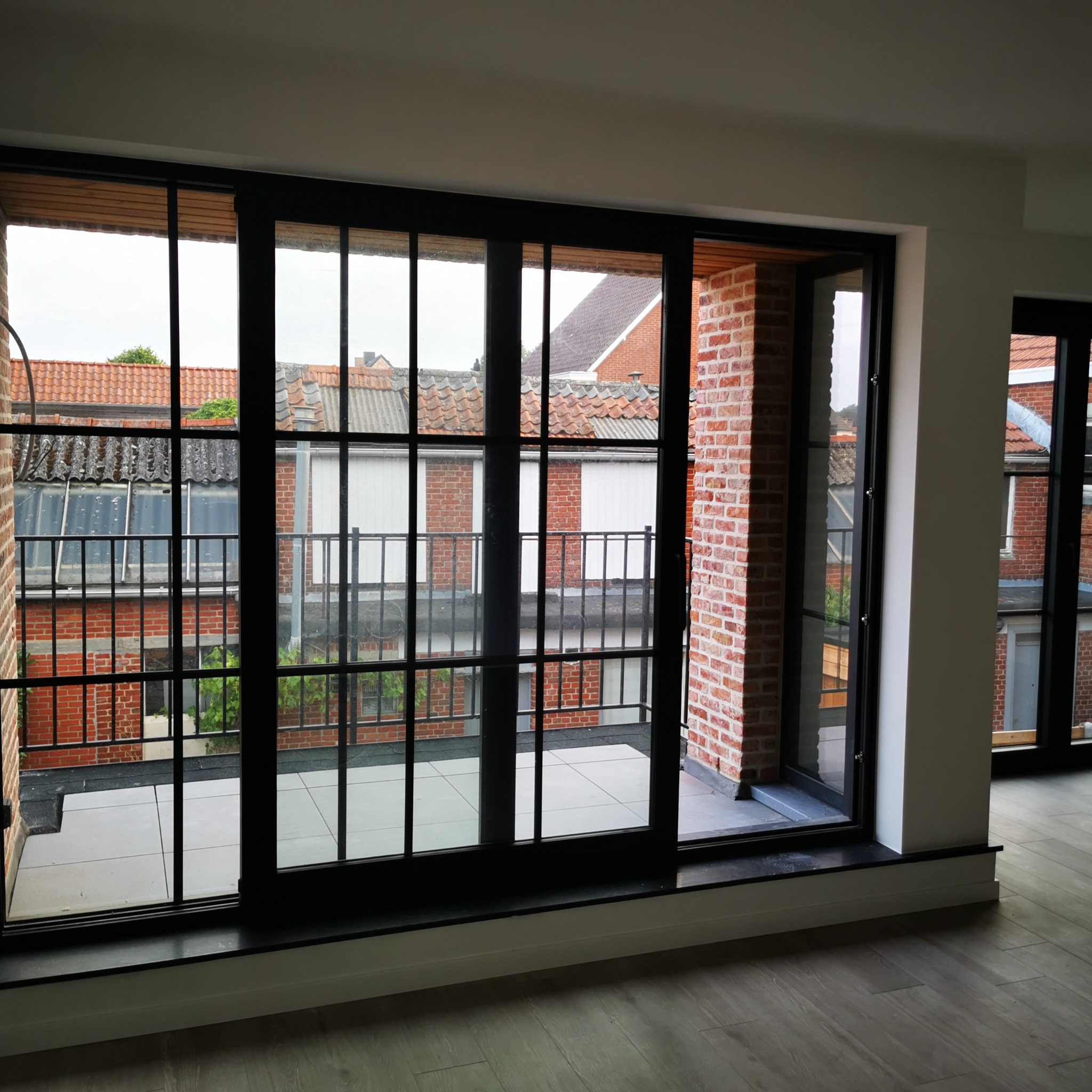 Nowoczesne, przeszklone drzwi balkonowe z czarnymi szprosami, widok na ceglane budynki z zewnątrz, jasna podłoga wewnątrz pomieszczenia.