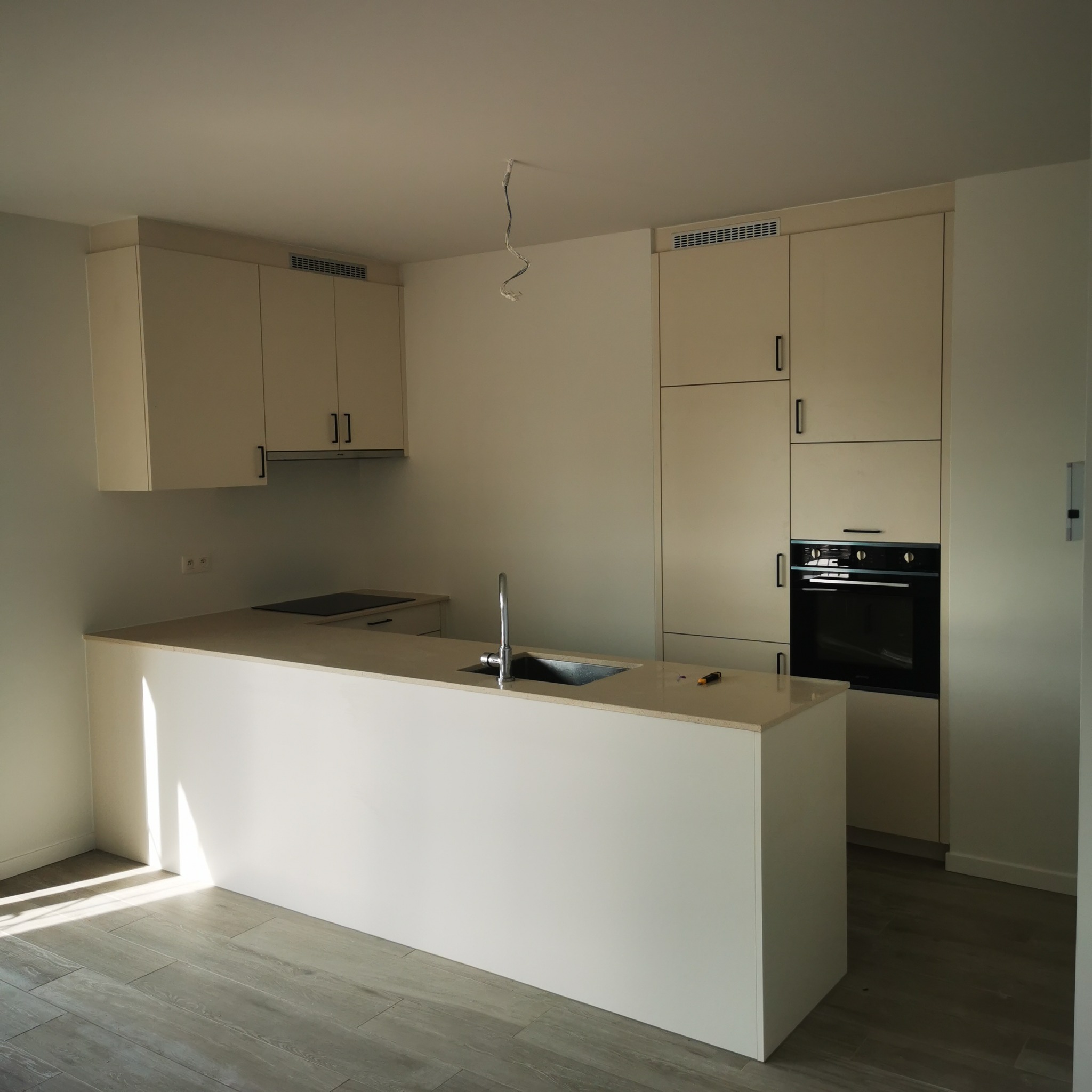 Nowoczesna, jasna kuchnia z wyspą i wbudowanymi szafkami, widoczne niedokończone podłączenie elektryczne do oświetlenia sufitowego.