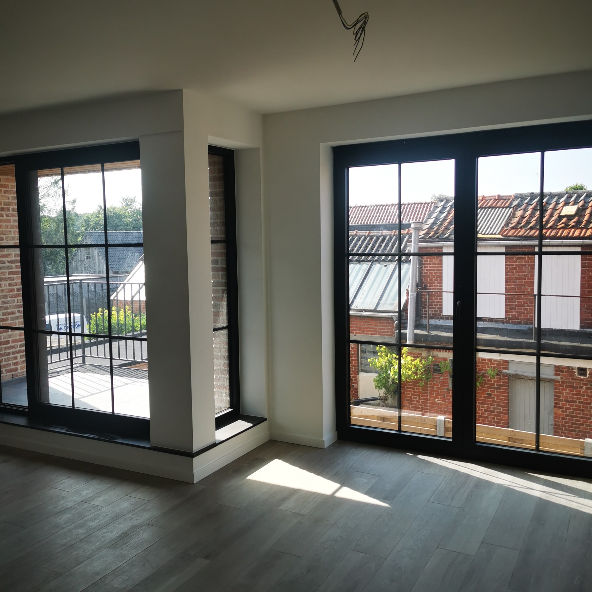 Jasne, przestronne wnętrze z dużymi oknami w czarnych ramach, widok na ceglany budynek z białymi okiennicami i zielenią, szare panele podłogowe ułożone w jodełkę.