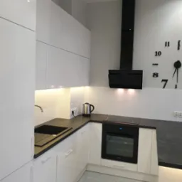 Nowoczesna, biała kuchnia z czarnym blatem, piekarnikiem i okapem. Na ścianie dekoracyjny zegar z cyframi.