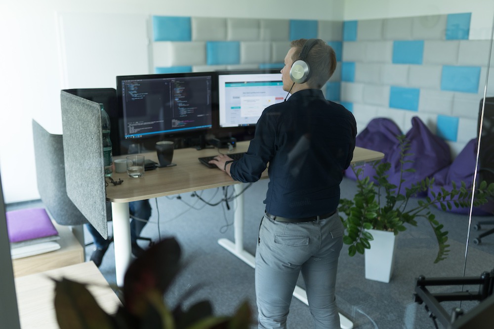 Programista w słuchawkach pracuje przy biurku z dwoma monitorami, na których widoczny jest kod i strona internetowa; w tle pufy i roślina doniczkowa.