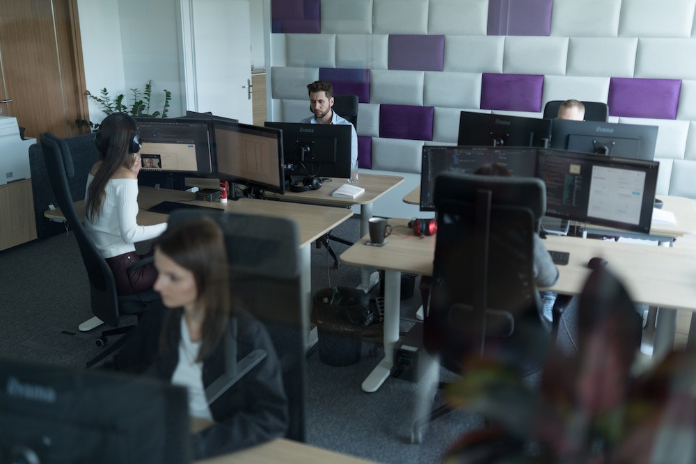 Wnętrze biura IT z pracownikami przy biurkach wyposażonych w monitory; jeden z pracowników prowadzi wideokonferencję.