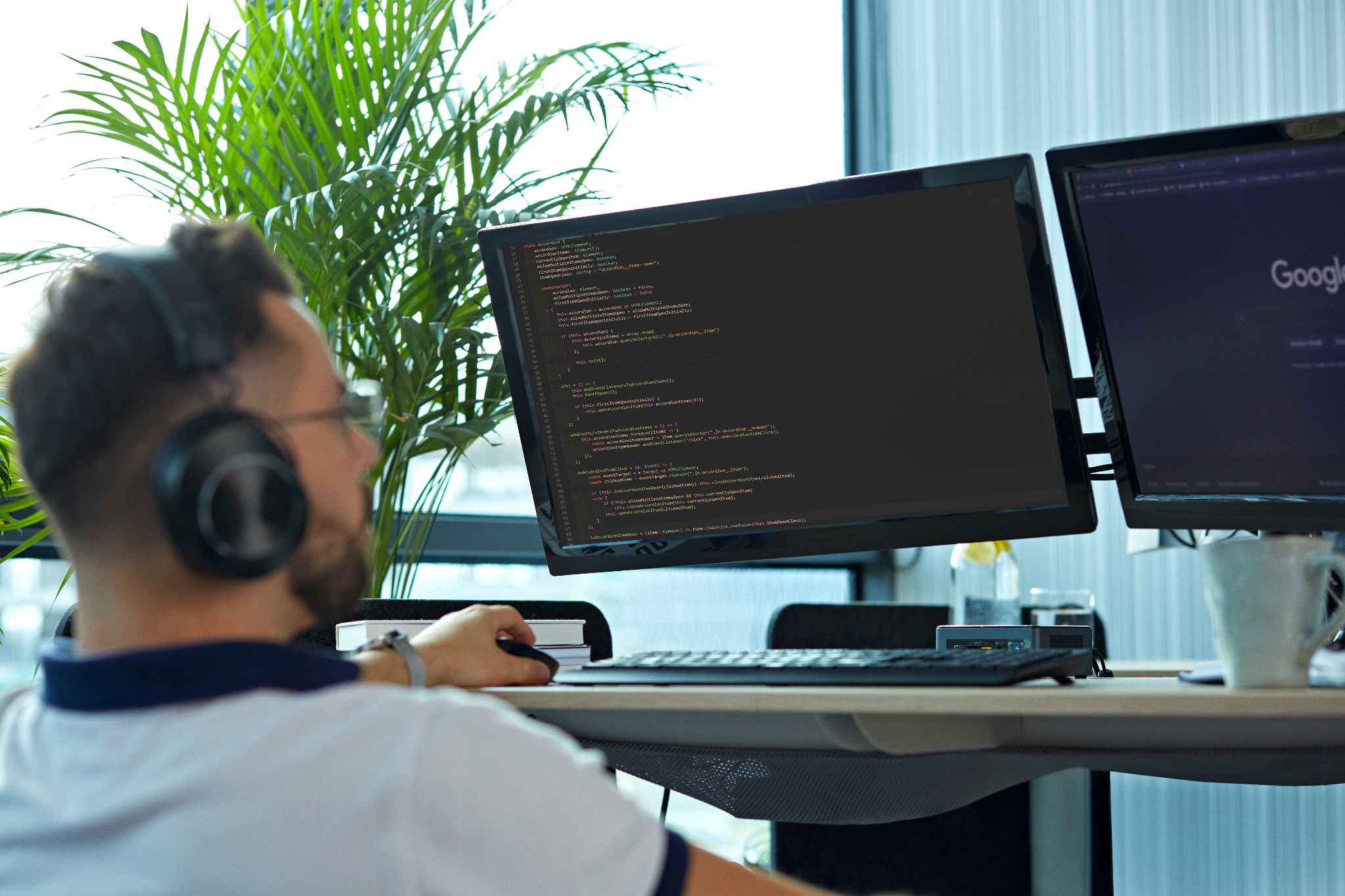 Programista w okularach i słuchawkach pracuje przy biurku z dwoma monitorami wyświetlającymi kod, z rośliną w tle i kubkiem po prawej stronie.