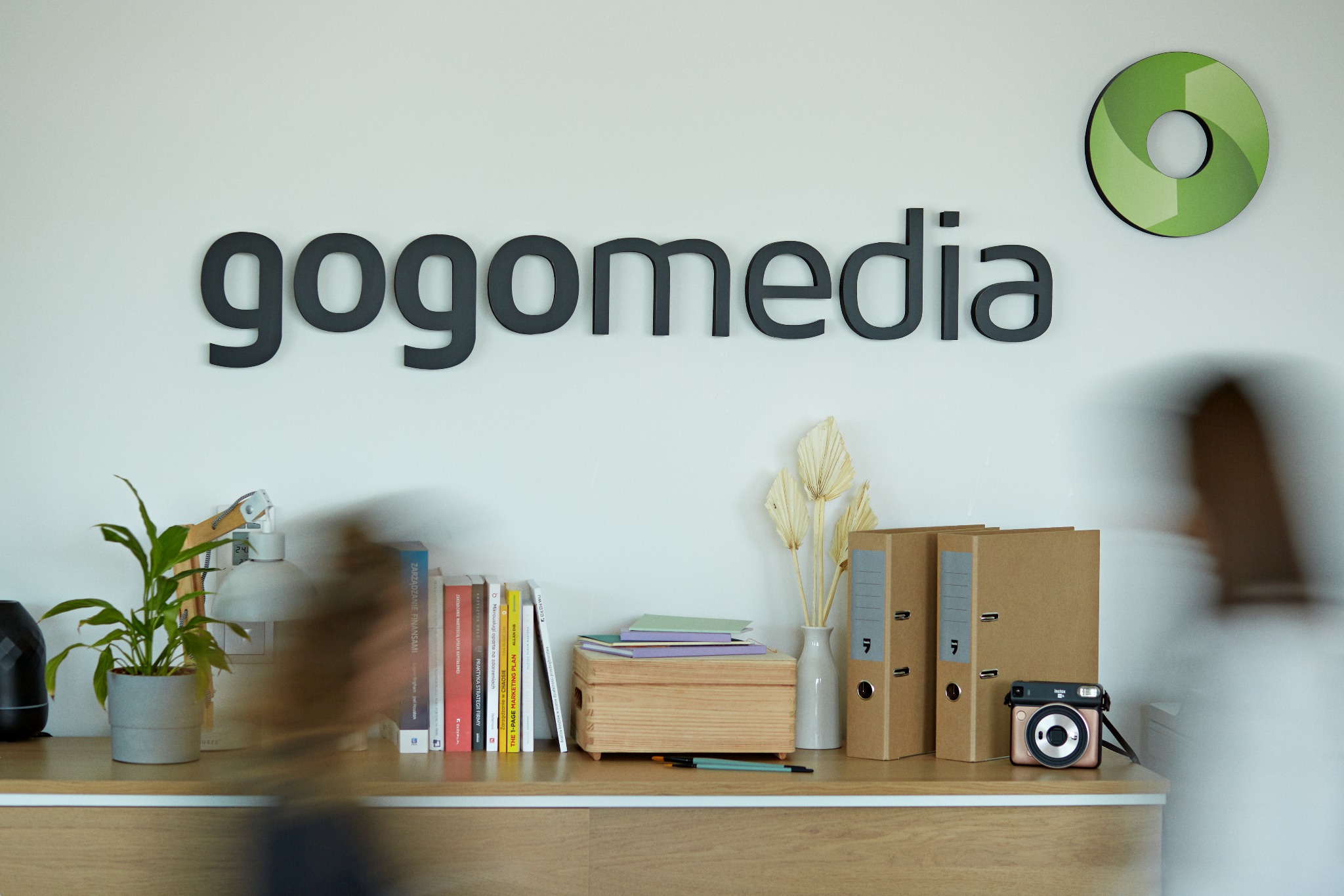 Biuro agencji marketingowej: logo 'gogomedia' na ścianie, roślina doniczkowa, książki, dokumenty w segregatorach, aparat Instax i dekoracje na drewnianej półce.