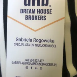 Wizytówka firmy Dream House Brokers, specjalisty ds. nieruchomości Gabriela Rogowska, z logo i danymi kontaktowymi na tle czarno-białej tkaniny.