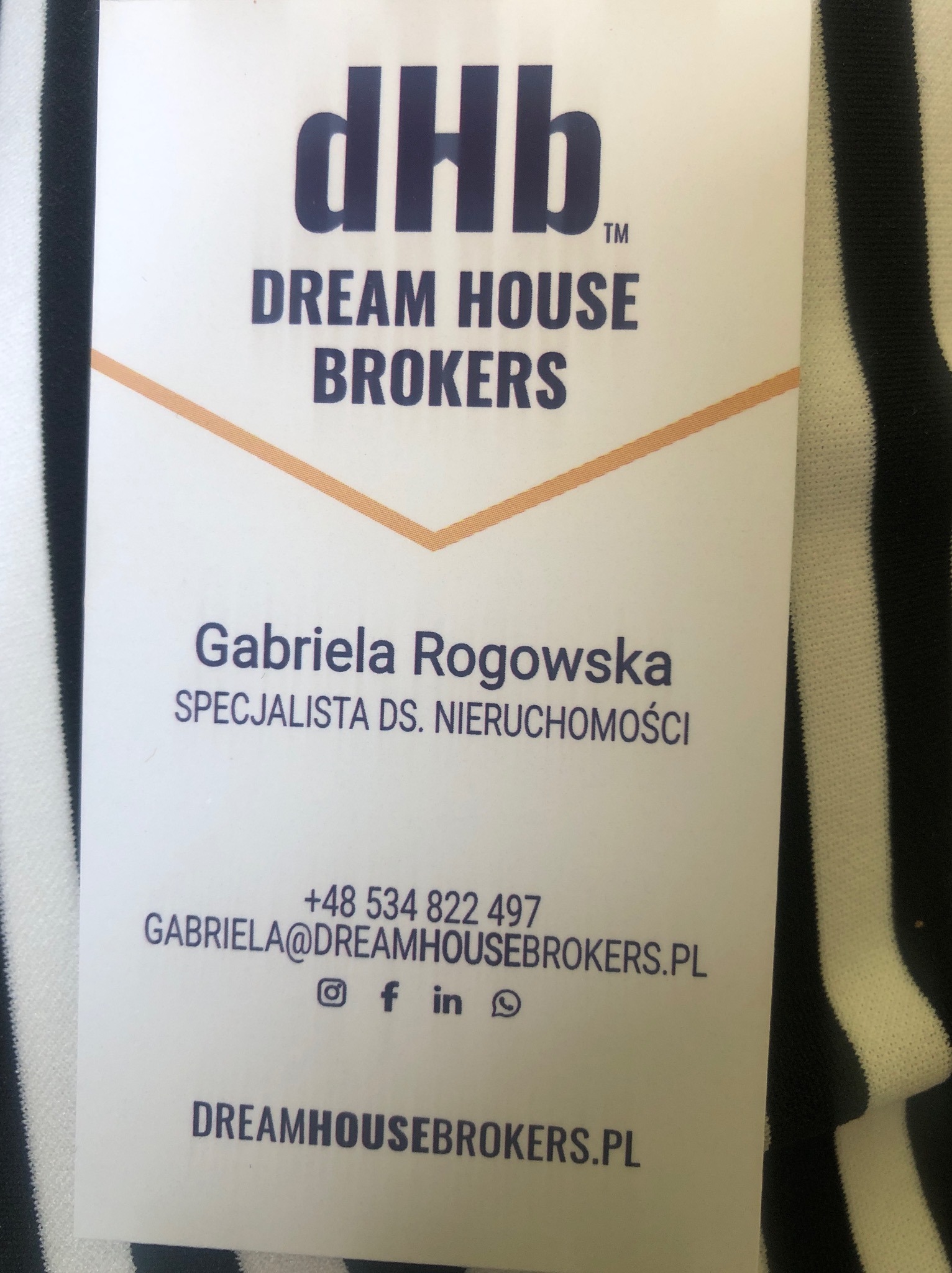 Wizytówka firmy Dream House Brokers, specjalisty ds. nieruchomości Gabriela Rogowska, z logo i danymi kontaktowymi na tle czarno-białej tkaniny.