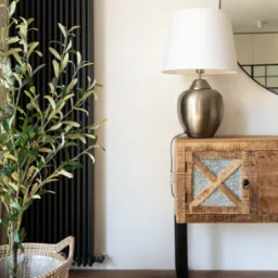 Stylowe wnętrze: drewniana komoda z lampą, okrągłe lustro, czarny grzejnik i roślina w koszu, tworzące przytulną aranżację w jasnym pomieszczeniu.