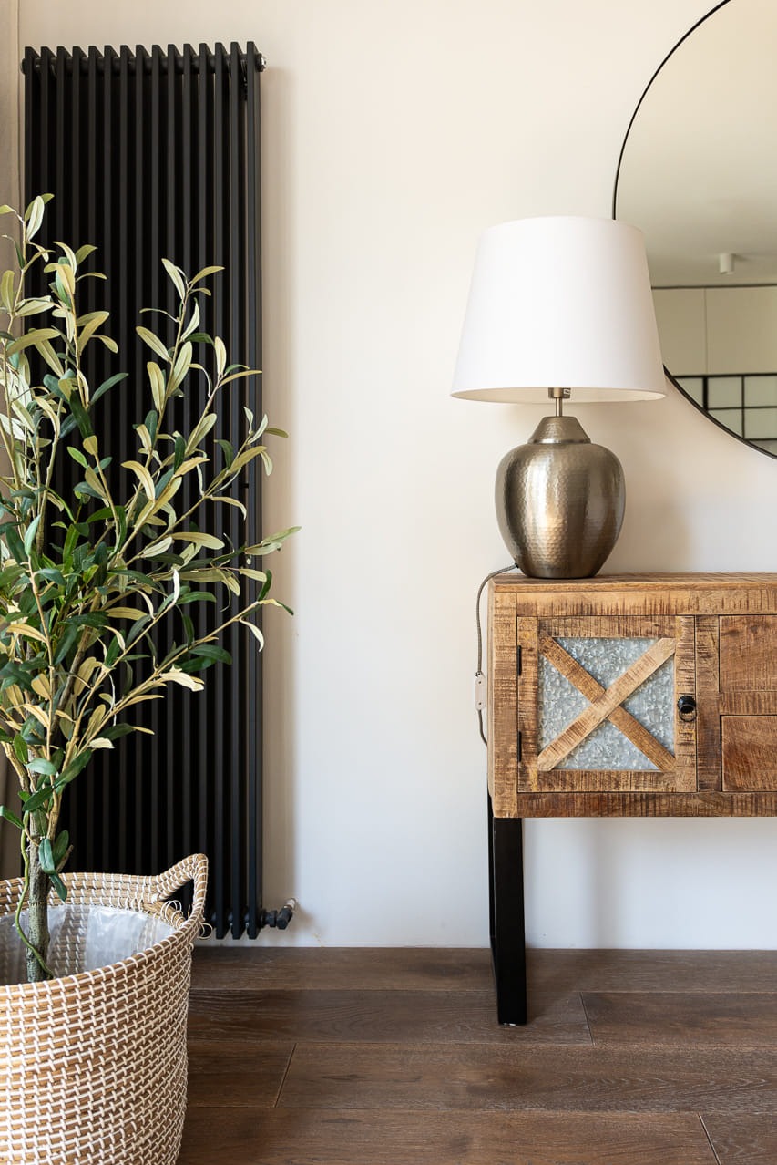 Stylowe wnętrze: drewniana komoda z lampą, okrągłe lustro, czarny grzejnik i roślina w koszu, tworzące przytulną aranżację w jasnym pomieszczeniu.