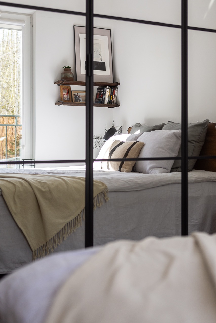 Jasna sypialnia za czarną, metalową ramą. Widoczny zagłówek, poduszki, pled i półka z dekoracjami. Za oknem zieleń. Minimalistyczny wystrój.