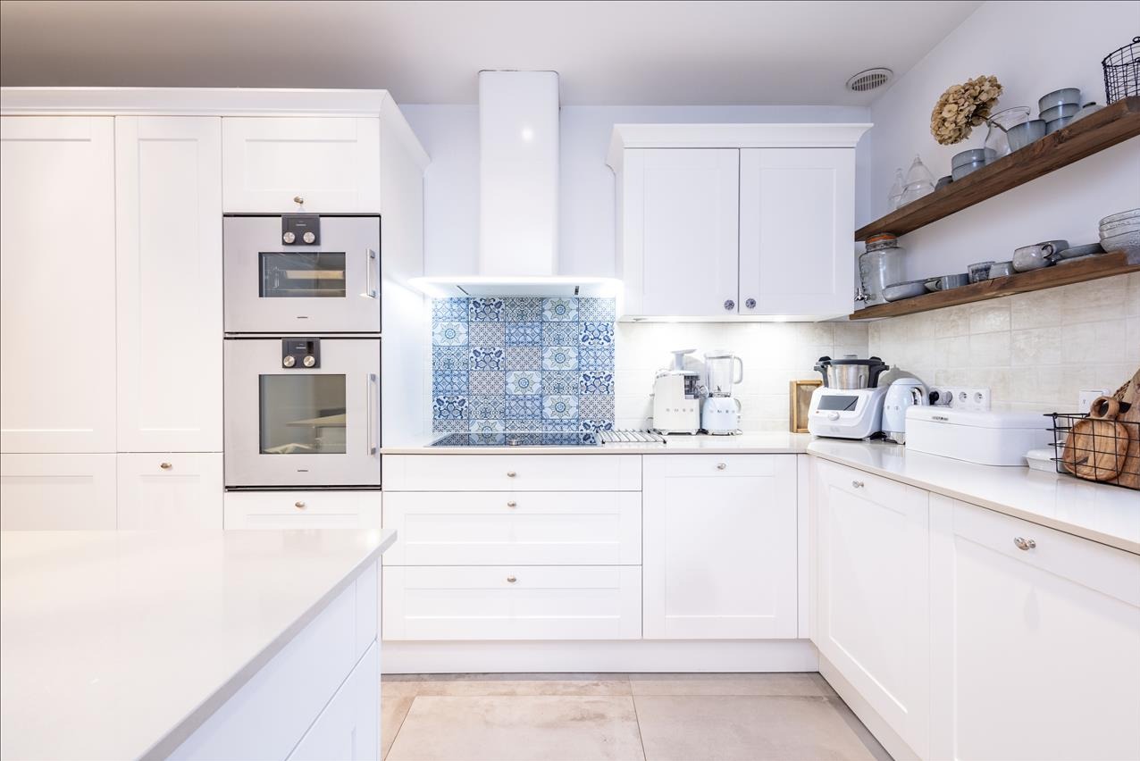 Jasna, nowoczesna kuchnia z białymi szafkami, sprzętem AGD marki SMEG i drewnianymi półkami z dekoracjami. Nad indukcją dekoracyjne, niebieskie płytki.