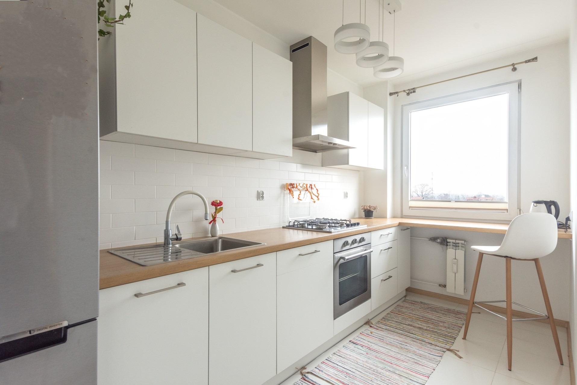 Jasna kuchnia w stylu skandynawskim: białe szafki, drewniany blat, stalowy okap i sprzęt AGD. Minimalistyczne oświetlenie i barowy stołek przy oknie.
