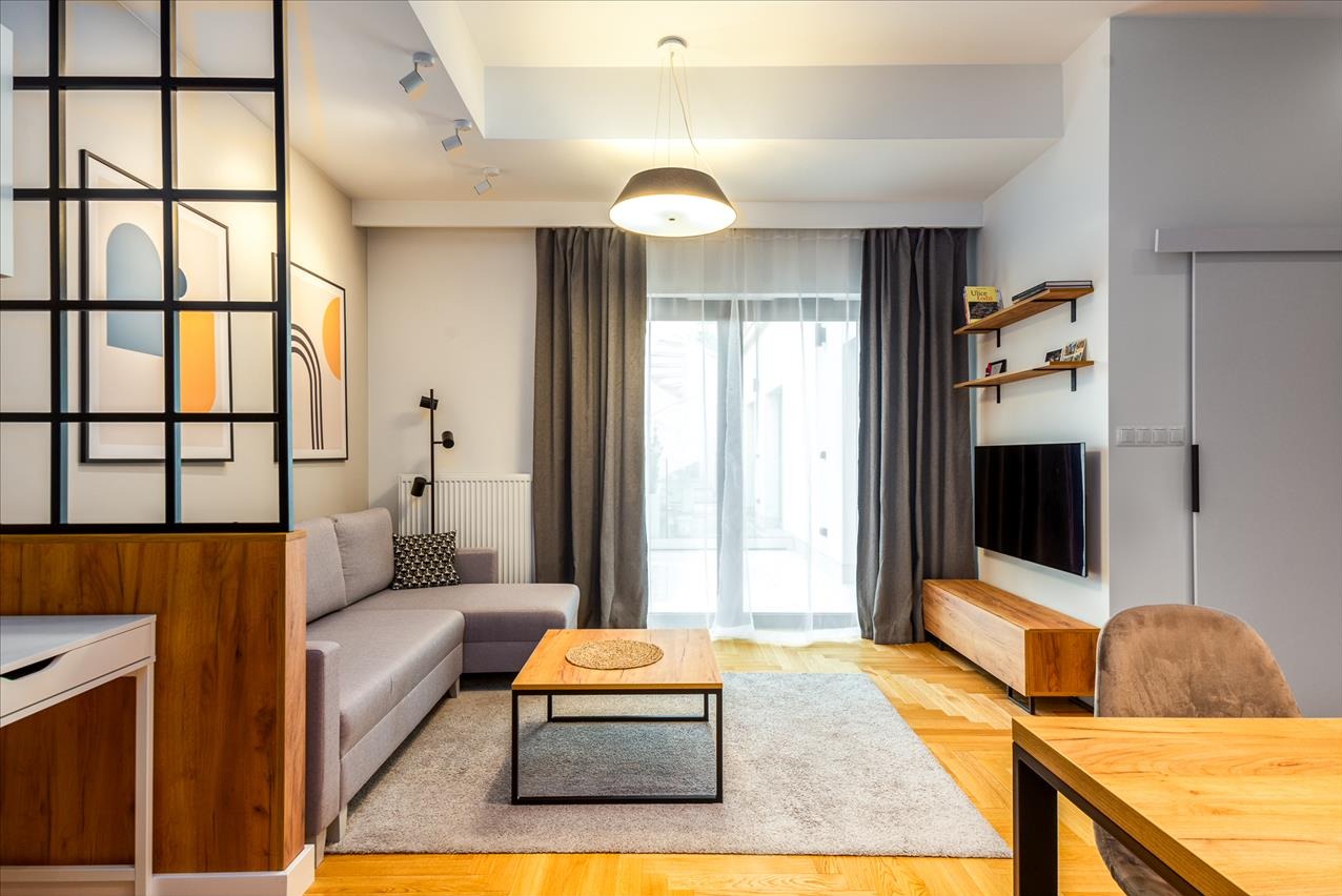 Jasne wnętrze mieszkania z szarą sofą narożną, drewnianym stolikiem kawowym na dywanie i dekoracjami ściennymi. Widoczne drewniane podłogi i naturalne światło.