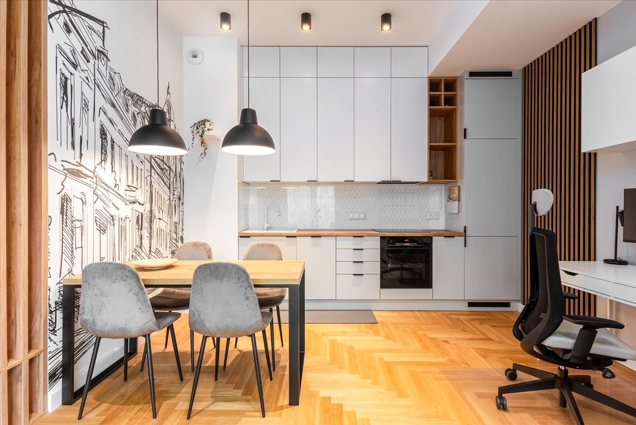 Jasne wnętrze mieszkania: drewniana podłoga, stół z krzesłami, biała zabudowa kuchenna, dekoracyjna ściana z motywem miasta i biurko z krzesłem obrotowym.