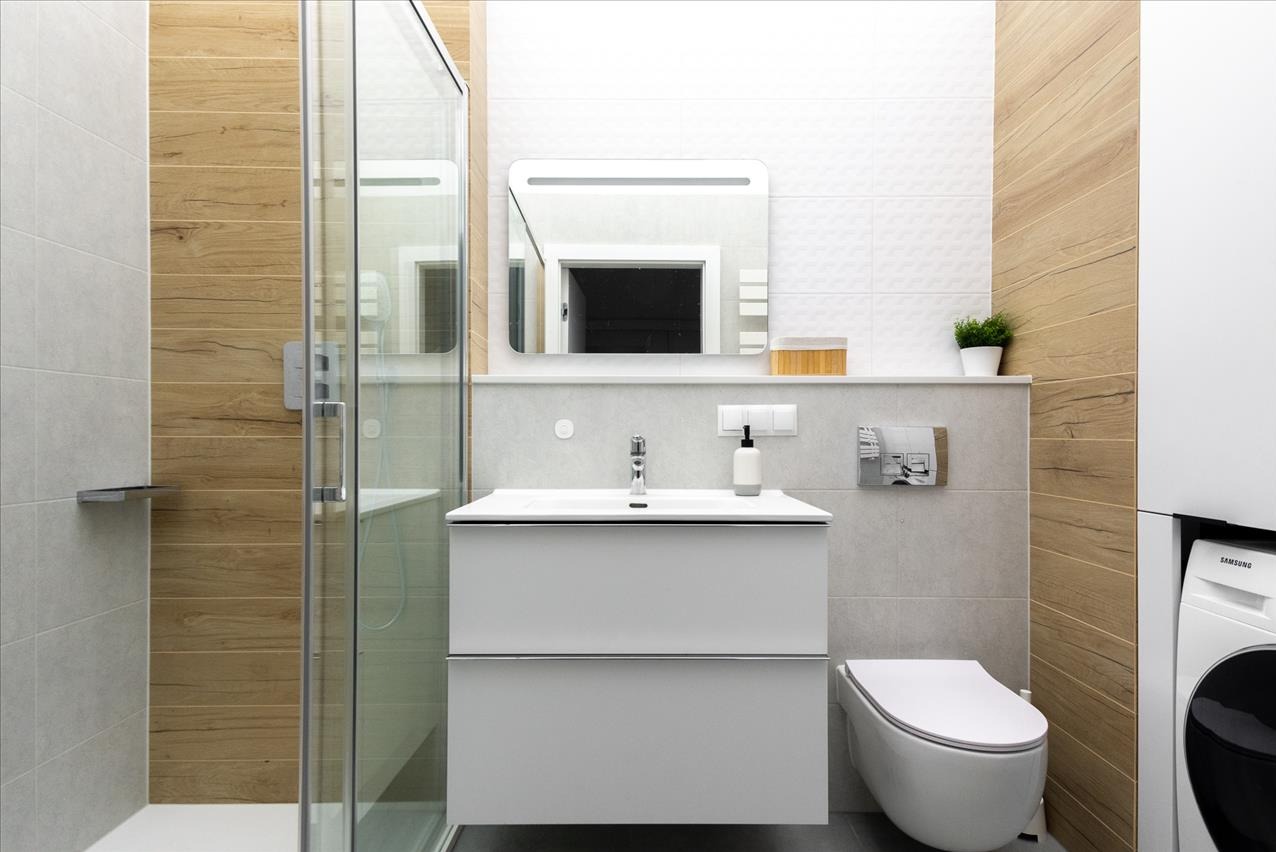 Nowoczesna łazienka z drewnopodobnymi płytkami, kabiną prysznicową i podświetlanym lustrem. Umywalka z szafką i toaleta w minimalistycznym stylu.