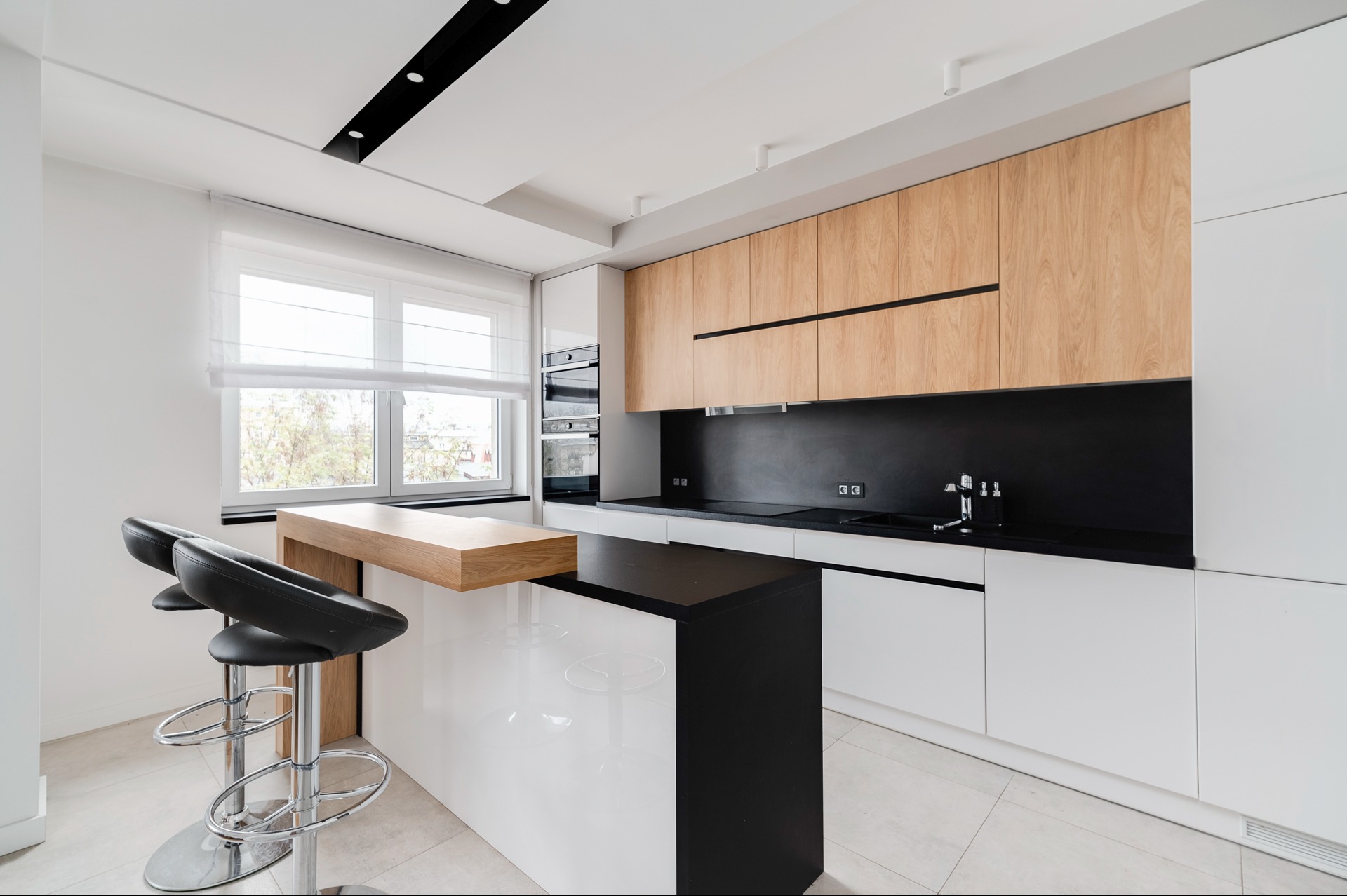 Nowoczesna kuchnia z wyspą i hokerami. Białe fronty szafek dolnych kontrastują z drewnianymi górnymi. Czarny blat i ściana nad blatem dodają elegancji.