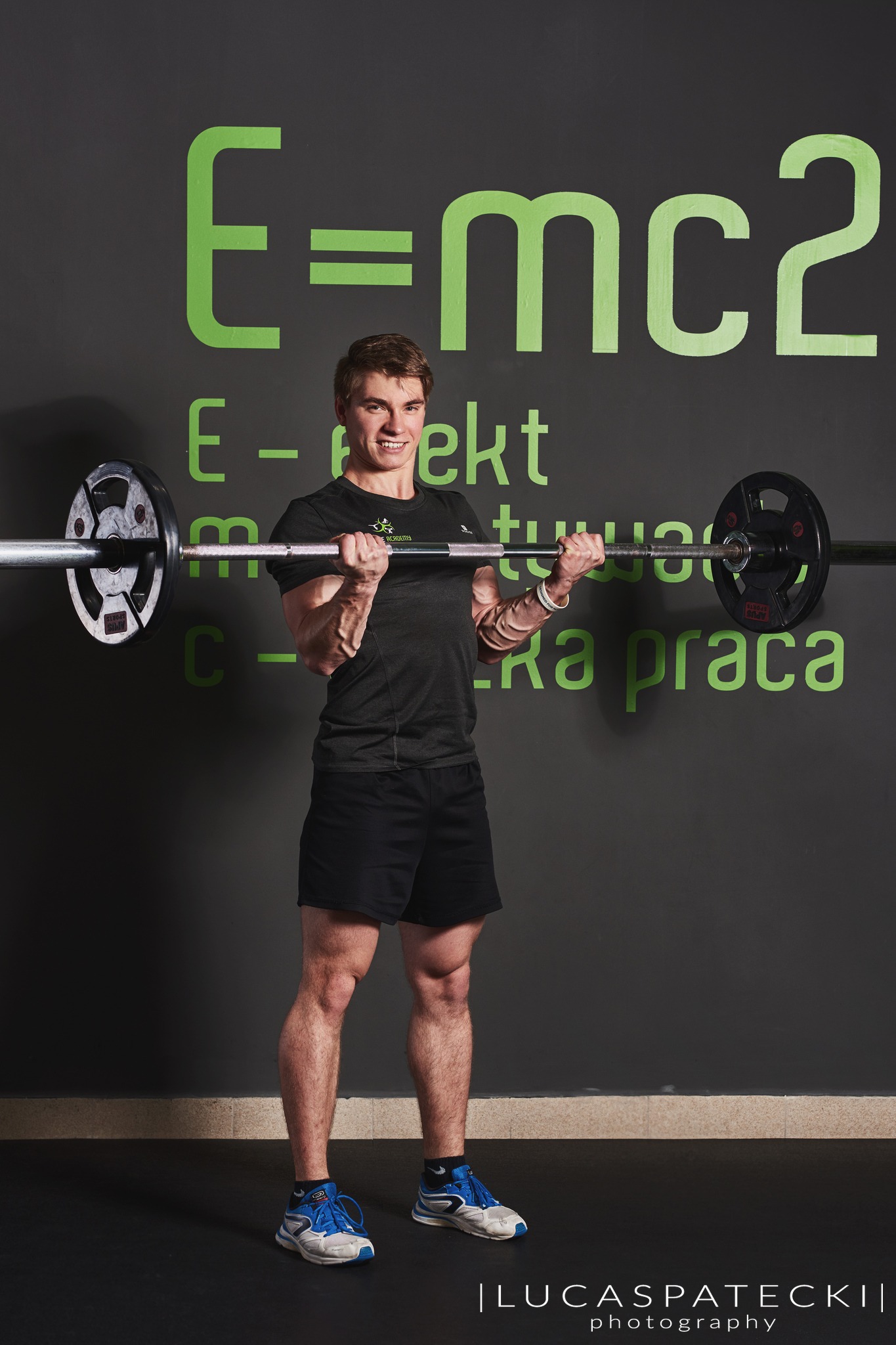 Młody mężczyzna w Krakowie ćwiczy biceps ze sztangą w siłowni z napisem E=mc2 na ścianie.