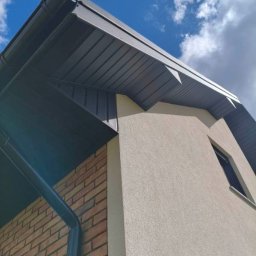 Usługi blacharskie - Szary dach z rynnami na budynku z ceglaną i otynkowaną ścianą, widok z dołu na tle błękitnego nieba z chmurami. Detal wykończenia dachu.