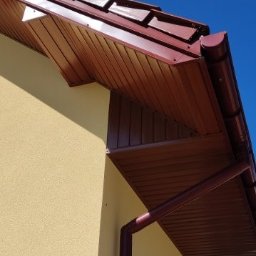 Usługi blacharskie - Ciemnoczerwony dach z rynnami i podbitką w kolorze brązowym na tle błękitnego nieba. Widok z dołu na fragment budynku z żółtą elewacją i oknem z roletami.