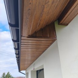 Usługi blacharskie - Podbitka dachowa z efektem drewna i czarna rynna na tle białej ściany budynku. Widok z dołu, podkreślający detale wykończenia i estetykę.