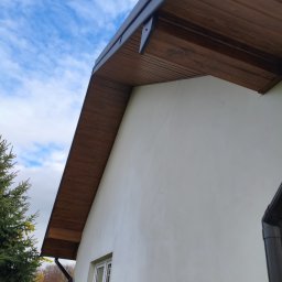 Usługi blacharskie - Drewniana podbitka dachowa z szarą rynną na tle białej ściany budynku i błękitnego nieba z chmurami. Widok z dołu, z boku budynku.