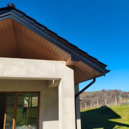 Usługi blacharskie - Nowoczesny dom z drewnianą podbitką dachową i czarnymi rynnami w słoneczny dzień. Widoczny fragment elewacji i okno z odbiciem osoby.