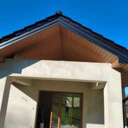 Usługi blacharskie - Nowoczesny dom z ciemnym dachem i drewnianą podbitką, widok z dołu na fragment elewacji i okno z odbiciem osoby. Błękitne niebo w tle.