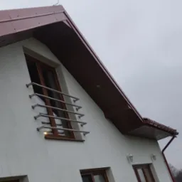 Dom jednorodzinny z nowym, brązowym dachem z blachy, brązowymi oknami i stalową balustradą okienną, sfotografowany w pochmurny dzień.