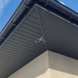 Niski kąt widzenia na narożnik dachu z czarną rynną i szarą blachą trapezową, przymocowaną do ściany budynku w słoneczny dzień. Biały sznurek zwisa z dachu.