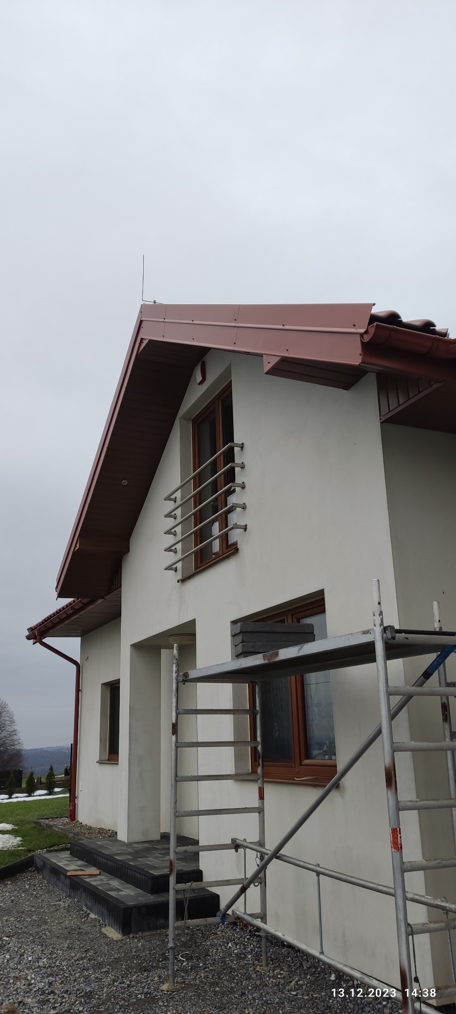Dom jednorodzinny z nowym, brązowym dachem z blachy, widoczna rynna w kolorze dachu, metalowe rusztowanie przy ścianie, świeżo ułożone schody z kostki brukowej prowadzące do wejścia, pochmurne...