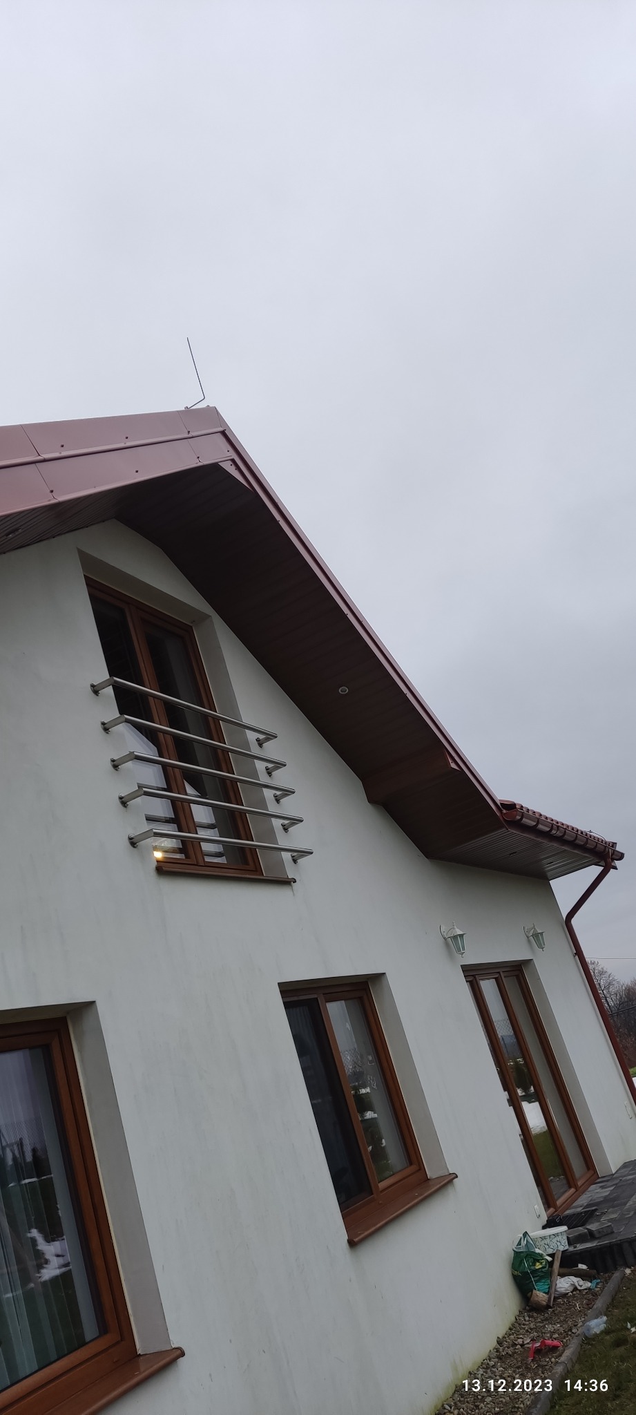 Dom jednorodzinny z nowym, brązowym dachem z blachy, brązowymi oknami i stalową balustradą okienną, sfotografowany w pochmurny dzień.