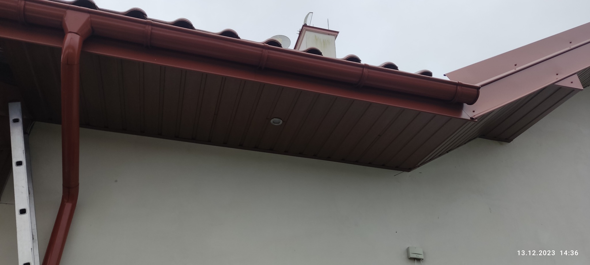 Brązowe rynny zamontowane na budynku z widoczną drabiną opartą o ścianę, dachówka w tle i komin z antenami.