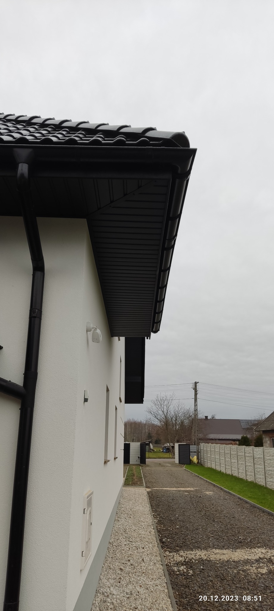 Fragment domu z czarnym dachem z dachówki ceramicznej, widoczna rynna i podbitka w kolorze czarnym, elewacja biała, obok podjazd wyłożony drobnym kamieniem.