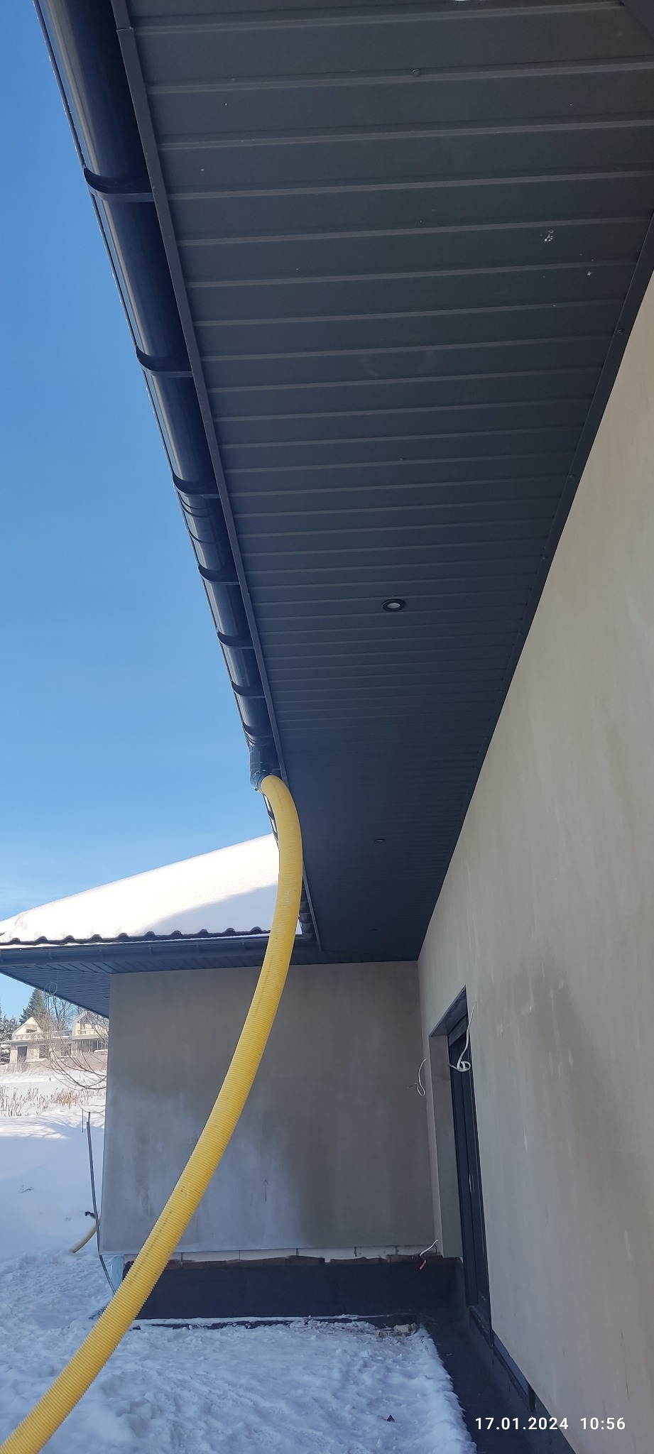 Nowy, grafitowy dach z blachy trapezowej i system rynnowy z żółtą rurą spustową odprowadzającą wodę, widok z dołu w zimowy dzień.